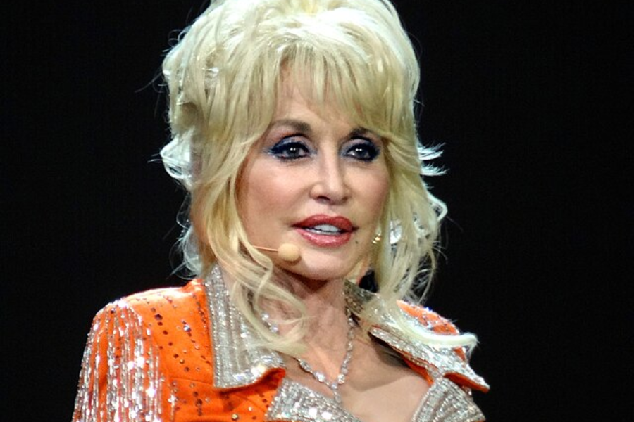 Blonde woman with voluminous hair and sparkly orange jacket speaks on stage wearing a headset microphone.