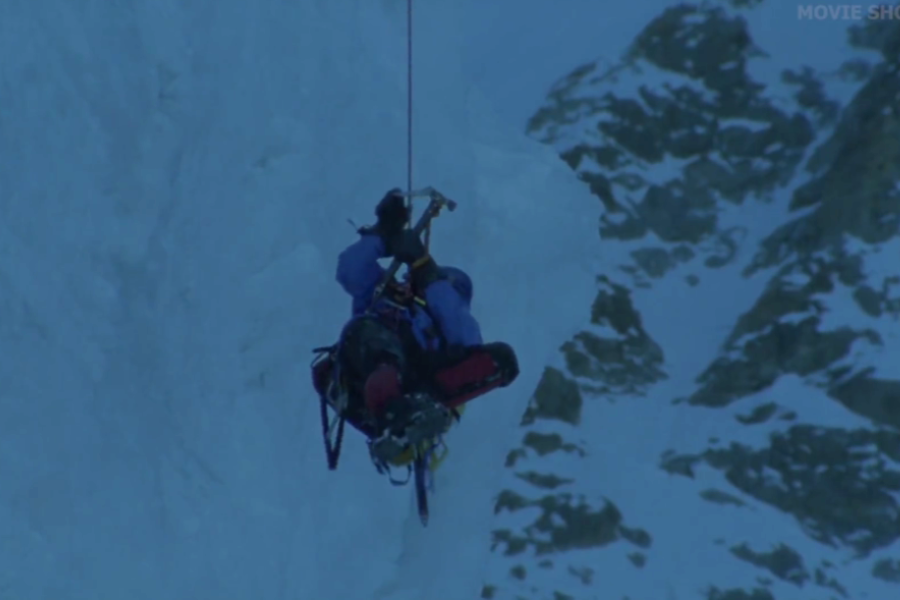 A mountaineer hangs mid-air, suspended by rope, as they assist another climber during a vertical descent on a steep, icy mountain face.