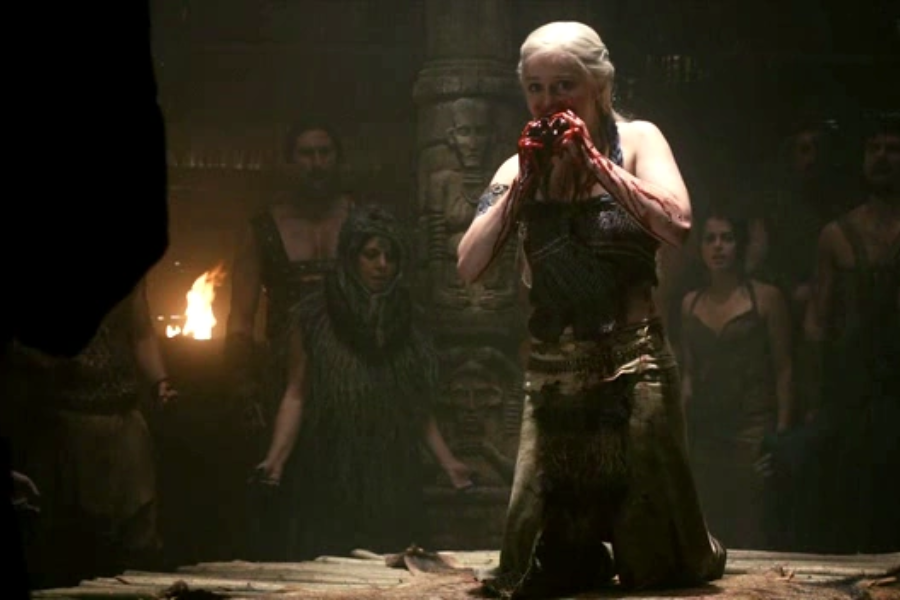 A woman with long white hair kneels in a dimly lit room, holding and eating a raw, bloody heart with onlookers behind her.