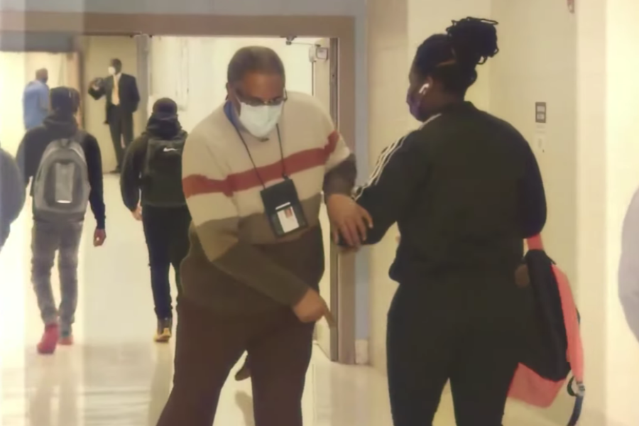 Man in a striped sweater and mask bumps elbows with a student in a school hallway.