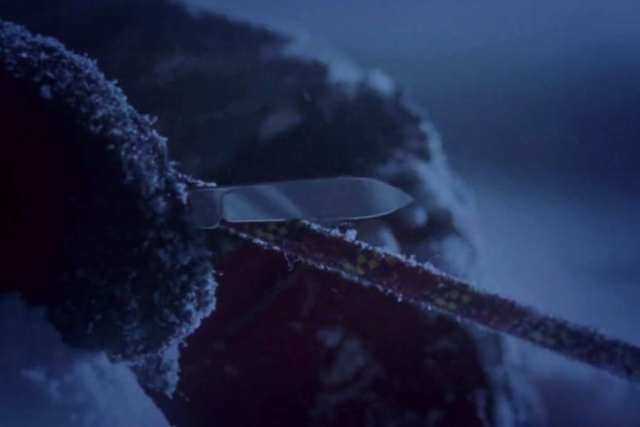 A frost-covered hand holds a knife against a rope in dim light, implying a critical moment of decision or survival in a cold environment.
