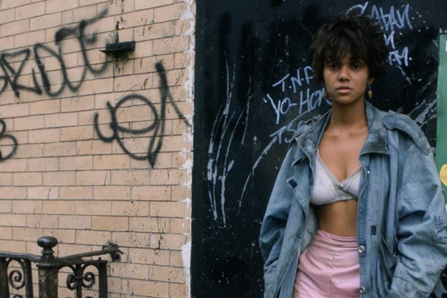 A young woman stands against a graffiti-covered wall wearing a large denim jacket, pink skirt, and exposed bra, staring seriously at the camera.