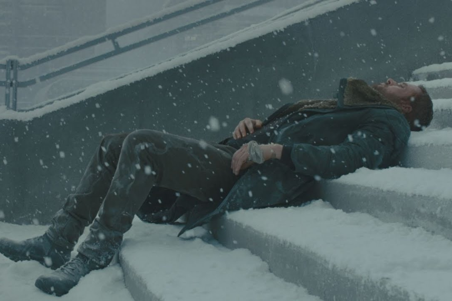 Man lying on snowy concrete stairs in heavy snowfall, appearing cold and motionless with his eyes closed.