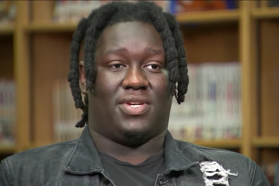Young man with short twists in a denim jacket speaks in a library setting.