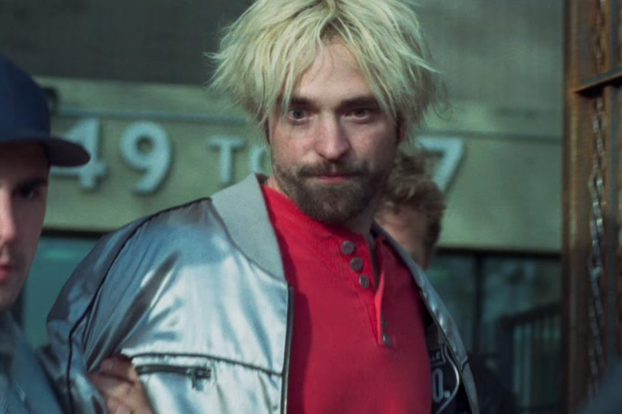 A disheveled man with bleached blonde hair and a beard wears a shiny silver jacket and red shirt while being escorted by police.