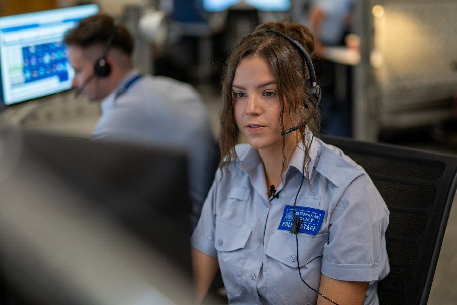 Female emergency dispatcher in uniform wearing a headset, speaking into a microphone at a call center.