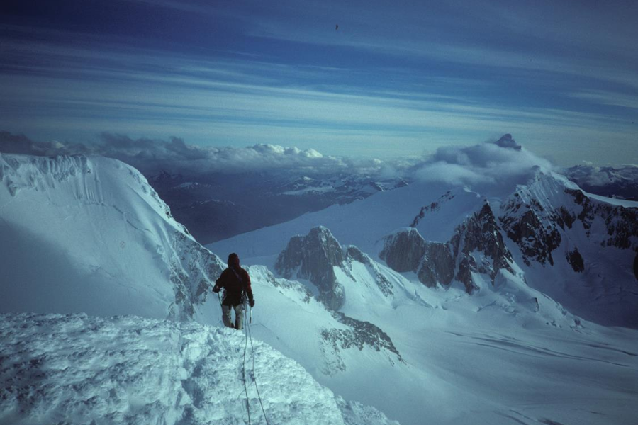 A lone mountaineer stands at the edge of a snow-covered ridge, overlooking a panoramic view of rugged, glacier-covered peaks and valleys under a moody sky.