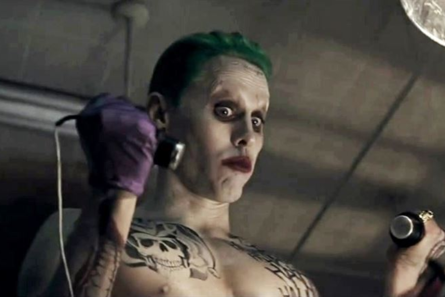 A shirtless man with pale skin, green slicked-back hair, and heavy makeup holds two electroshock paddles with a wide-eyed expression.