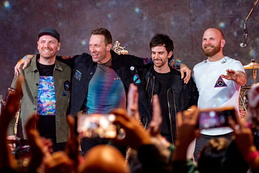 Four band members stand arm-in-arm on stage, smiling at a cheering crowd taking photos with smartphones.