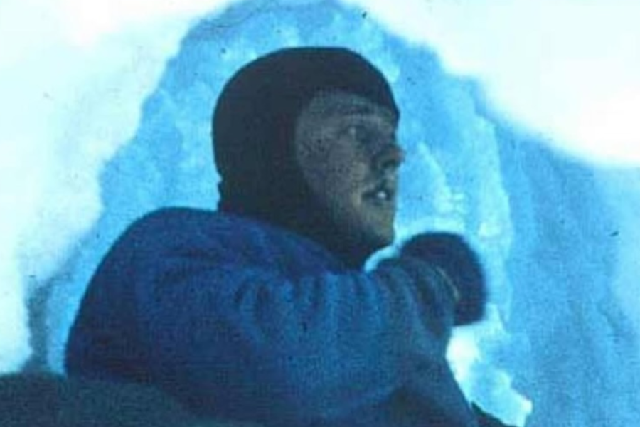 A man dressed in cold-weather gear is seen inside a narrow icy crevasse, surrounded by blue ice walls with light filtering from above.