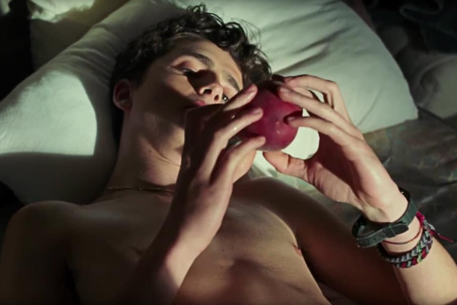 A shirtless young man lies in bed holding a peach, staring at it contemplatively in a softly lit bedroom.