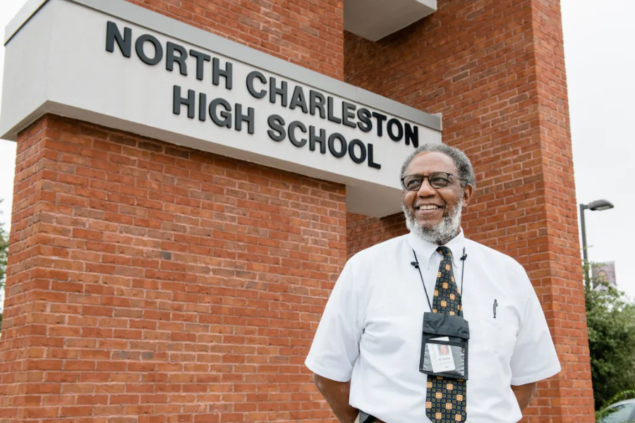  Smiling man in a white shirt and patterned tie stands in front of the North Charleston High School sign on a brick building.