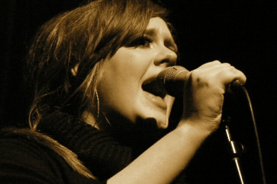 Close-up of a woman singing emotionally into a microphone in a sepia-toned photo, wearing a dark sweater.