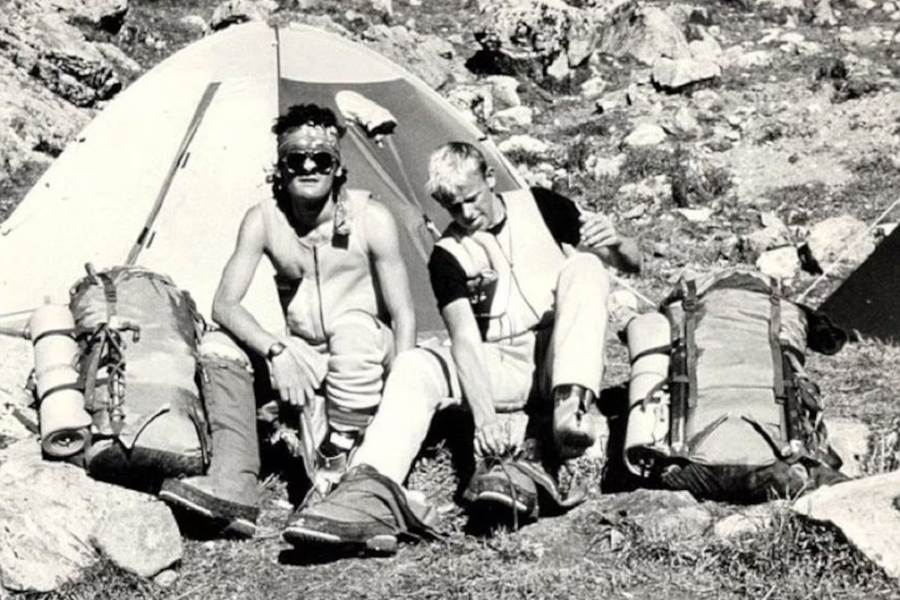 A vintage black-and-white photo shows two male climbers resting in front of a tent with heavy gear and backpacks, likely preparing for a high-altitude expedition.
