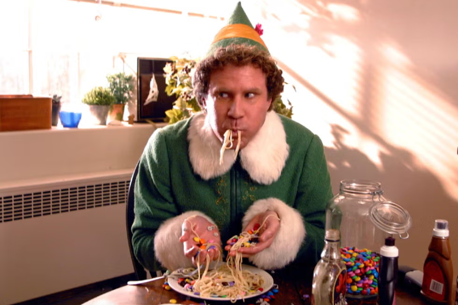 A man dressed as an elf sits at a table eating spaghetti with candy and syrup using his hands, looking playfully to the side.