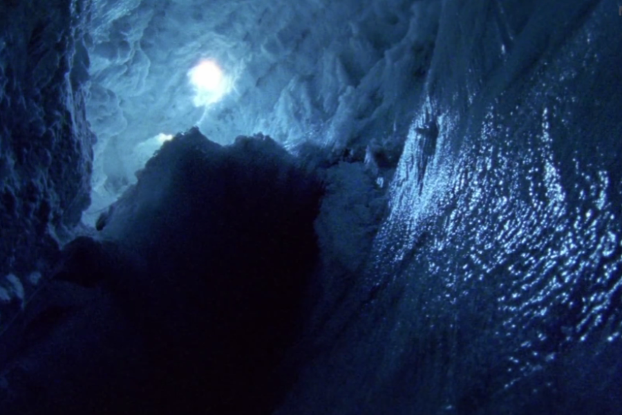 The view from inside a deep ice crevasse looking up toward the snowy opening, with glistening textured ice walls and faint light from above.