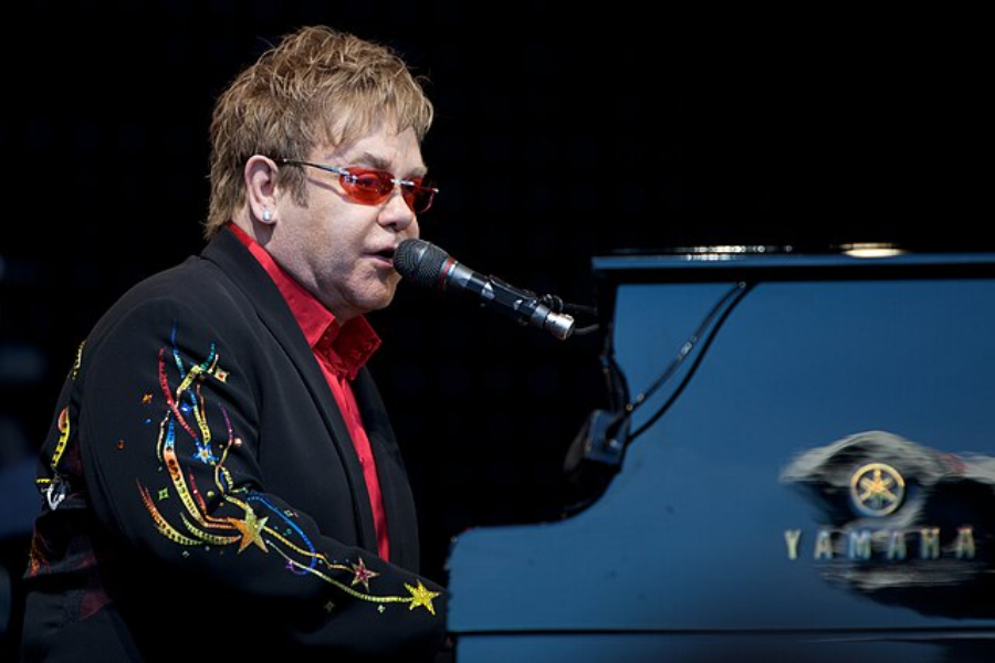 Man in red-tinted glasses and a black jacket with colorful embroidery performs while playing a Yamaha piano.