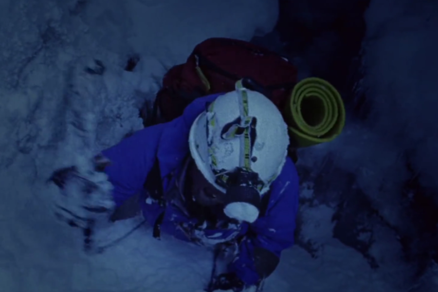 A climber in blue winter gear ascends through a narrow, snow-packed crevasse at night, wearing a helmet with a headlamp and carrying a red backpack with a rolled yellow foam mat.