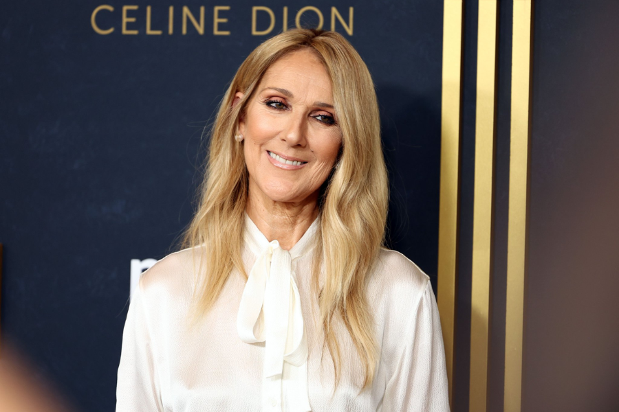 Woman with long blonde hair smiles softly while posing in a silky white blouse in front of a backdrop with her name.