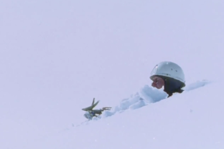 A climber’s head and ice axe barely protrude from deep snow, suggesting a fall into a crevasse or avalanche burial in a whiteout environment.