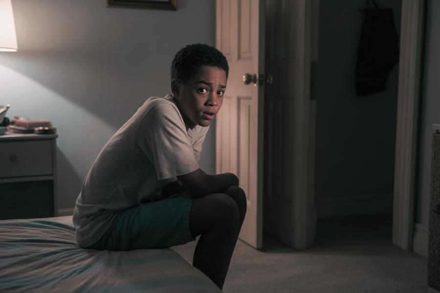 A young boy sits on the edge of a bed in a dimly lit bedroom, looking toward the doorway with a tense and anxious expression. The open door behind him and his body language suggest fear or worry about someone entering.
