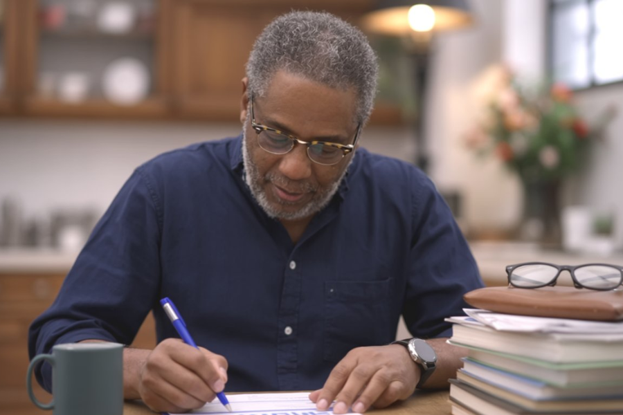 Man in a navy shirt writes on papers at a wooden table with a coffee mug and books nearby.