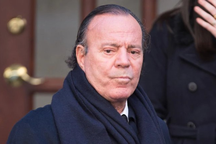 Older man wearing a dark coat and scarf stands outdoors with a serious expression.