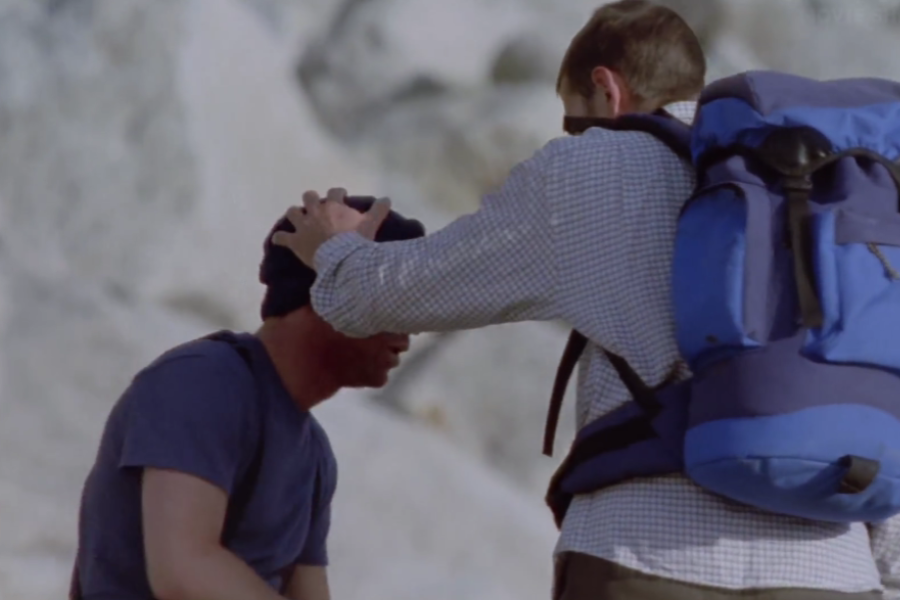 One man in a blue backpack gently touches the forehead of another seated climber, appearing to check on him or provide reassurance in a rocky alpine area.