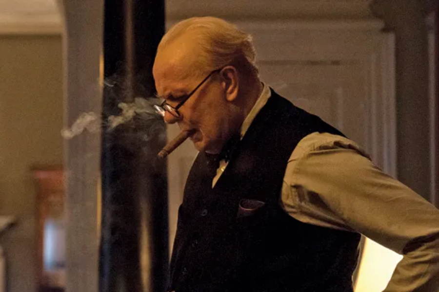 An older man in a vest and glasses smokes a cigar with smoke curling from his mouth in a dimly lit room.