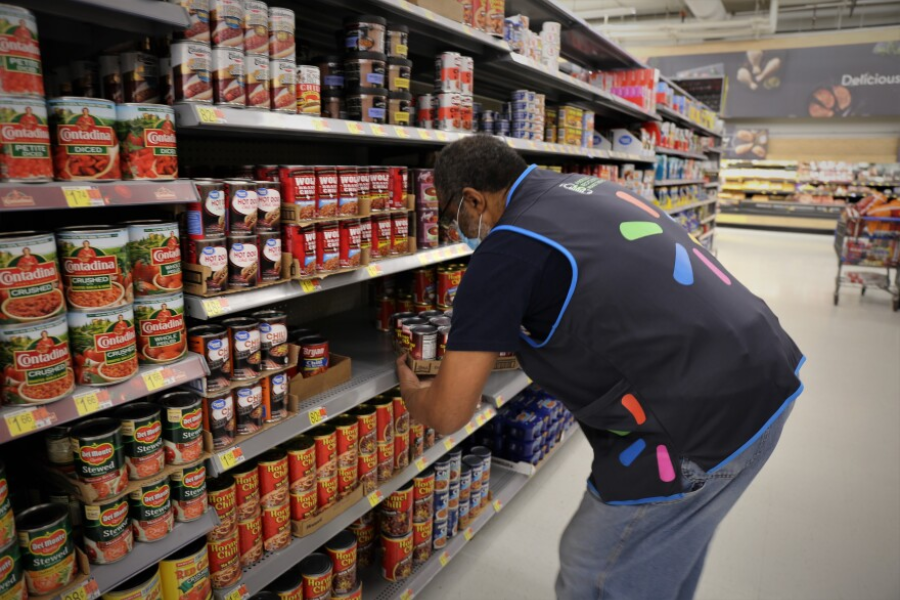 Walmart employee in a black vest stocks shelves with canned goods in the grocery aisle.