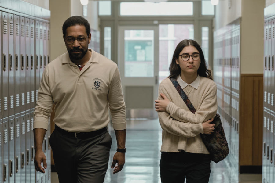 A teacher and a student walk side by side down a school hallway lined with lockers. The student looks anxious, crossing her arms, while the teacher walks with a serious expression.