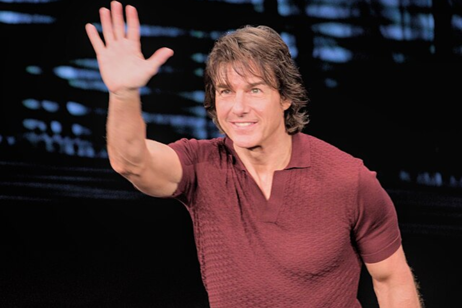 Man in a fitted maroon shirt smiles and waves with his right hand against a dark, blurry background.