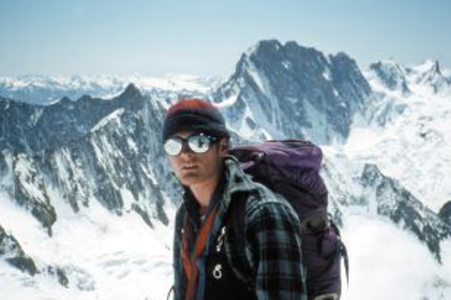 A climber wearing mirrored sunglasses and a dark headband poses with snow-covered peaks behind him, reflecting the mountain landscape in his lenses.