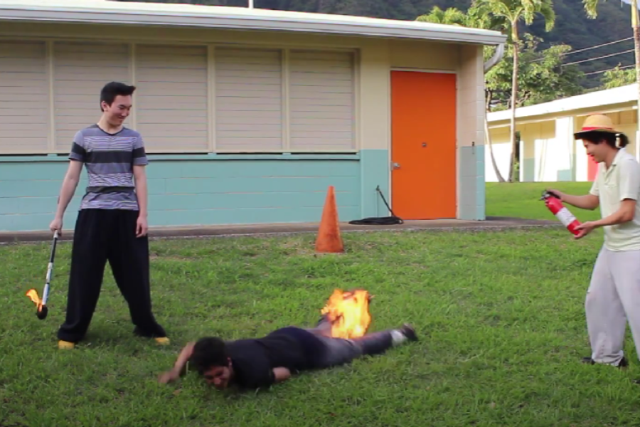 Person on fire rolling on grass while one individual holds a fire extinguisher and another stands nearby with a flaming baton.