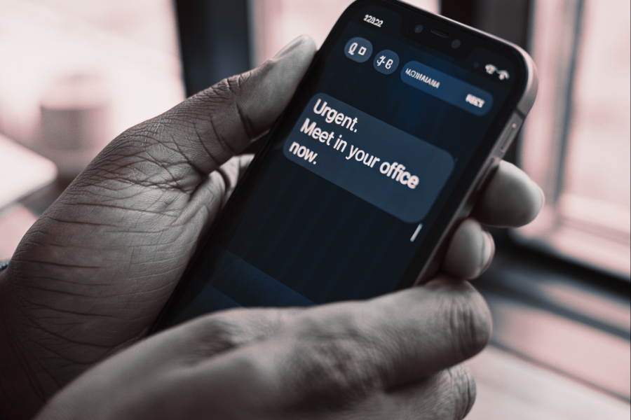 A close-up of a phone screen held in someone’s hands, showing a text message that reads “Urgent. Meet in your office now.” The message appears tense and immediate.