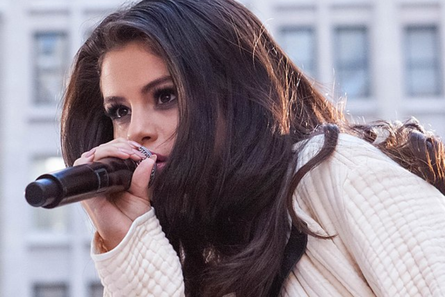 Young woman with voluminous dark hair sings into a microphone, wearing a textured white outfit in an outdoor setting.