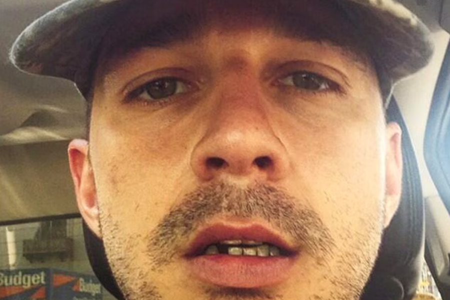 A man with a scruffy beard and grillz stares blankly into the camera from the front seat of a car.
