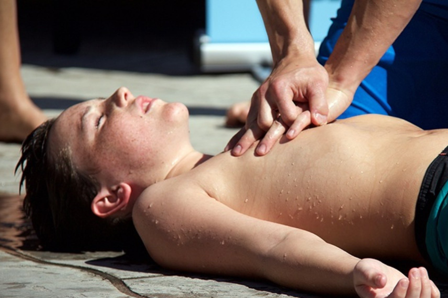 Close-up of someone performing chest compressions on a wet, unconscious child lying on a poolside surface.