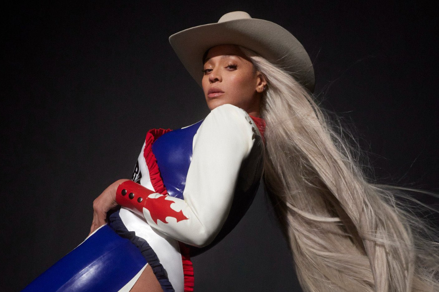Woman with long platinum hair poses confidently in a red, white, and blue outfit with a cowboy hat in dramatic lighting.