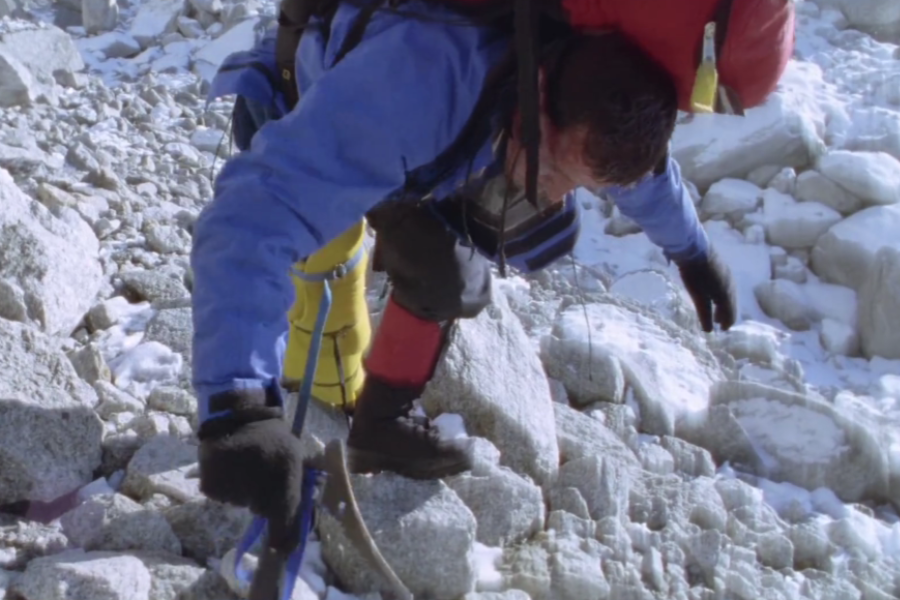 A climber in worn blue and yellow gear climbs over a rocky slope, appearing physically exhausted as he grips his ice axe and crawls forward.