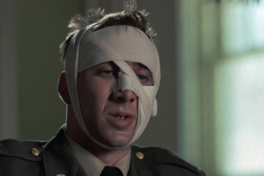 A man in military uniform sits with a bandaged face, looking weary and injured in a softly lit room.
