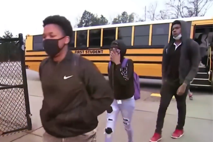 Students wearing masks walk away from a yellow school bus toward a school entrance.