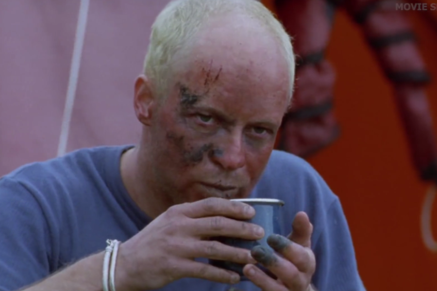 A man with a bruised and bloodied face drinks from a tin cup, his hands and face showing clear signs of injury and exposure.