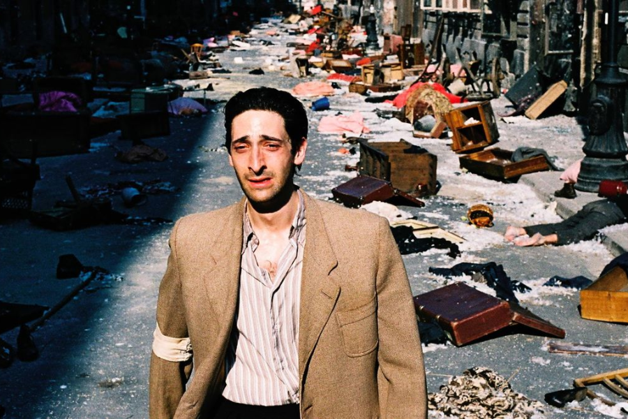 A man stands in a debris-strewn city street wearing a brown blazer and armband, squinting toward the sunlight.