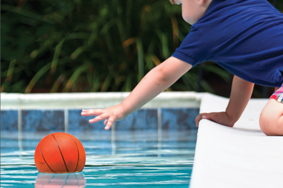 Child kneeling by the edge of a swimming pool, reaching out over the water to grab an orange basketball.