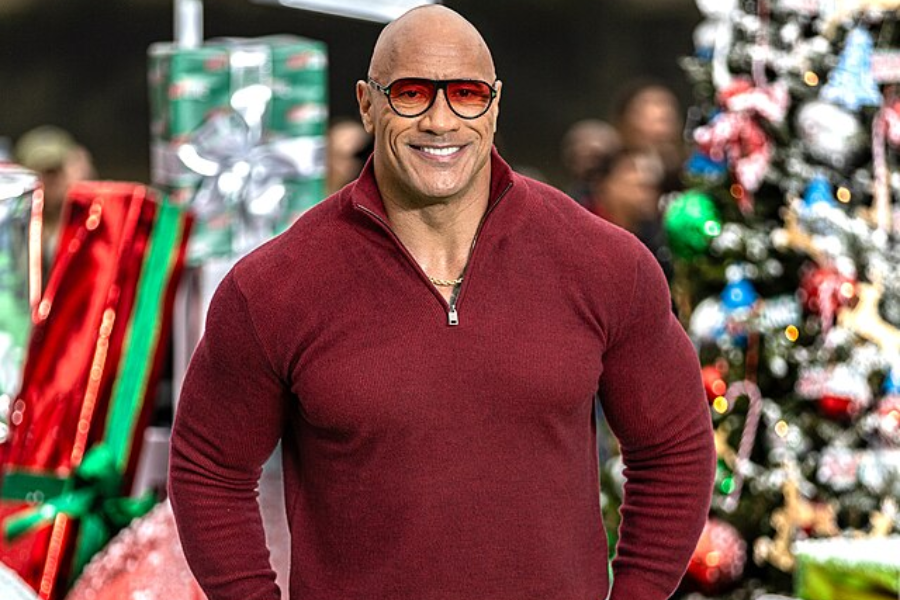 Muscular man in a red zip-up shirt smiles while standing in front of a Christmas tree and wrapped gifts.