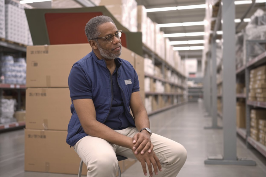 Walmart employee sits on a stool in a warehouse aisle surrounded by stacked cardboard boxes.