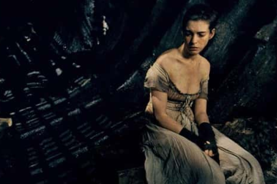 A woman in a tattered dress and fingerless gloves sits slouched and exhausted against a dark, grimy background.