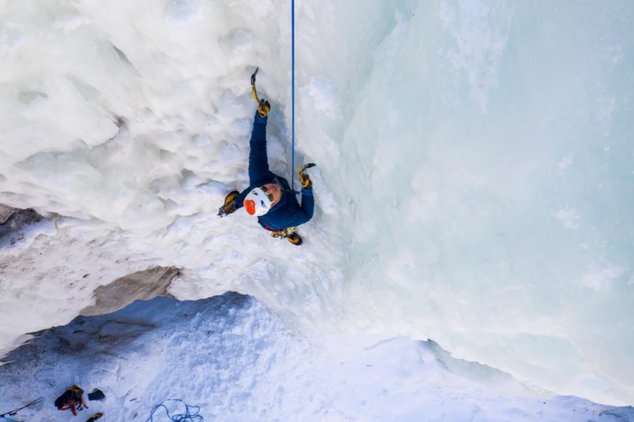 A climber in blue winter gear ascends a vertical ice wall using ice axes and a safety rope, surrounded by thick, textured ice formations.
