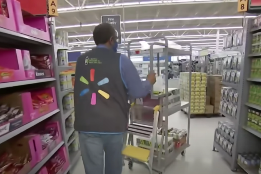 Walmart employee pushes a stocking cart down a grocery store aisle past shelves of candy and canned goods.